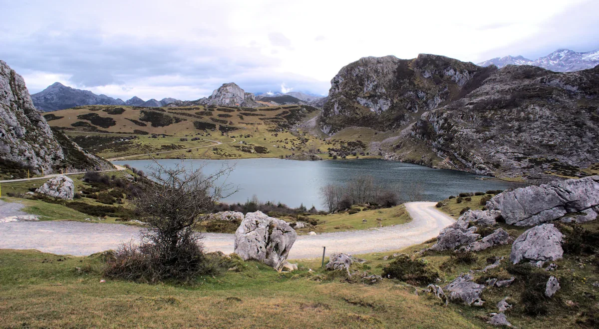 Lake Enol lies among rocky slopes and open grassland, with a winding road in the foreground and snowy peaks in the distance.
