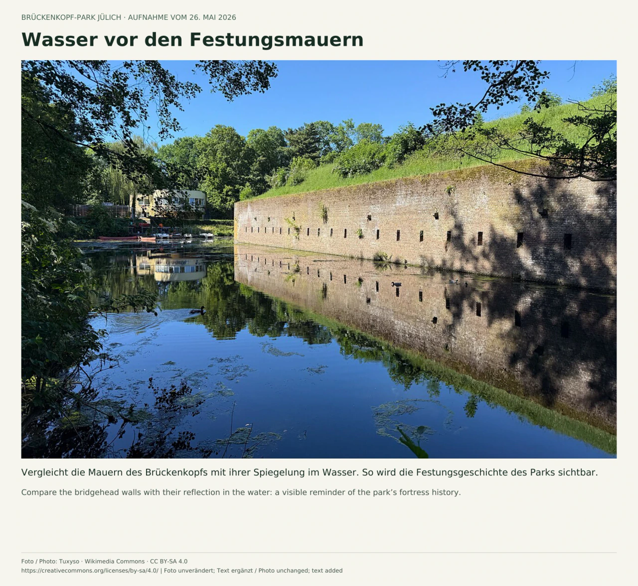Eine lange gemauerte Festungswand mit kleinen Öffnungen und grasbewachsenem Wall spiegelt sich im ruhigen Wassergraben; Bäume rahmen das Bild.