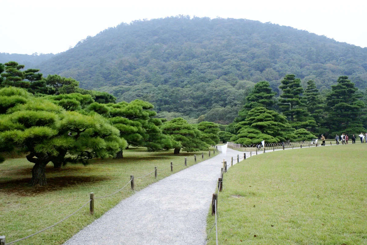 A gravel path curves between low sculpted pines and a lawn, with taller shaped trees and a forested hillside behind.