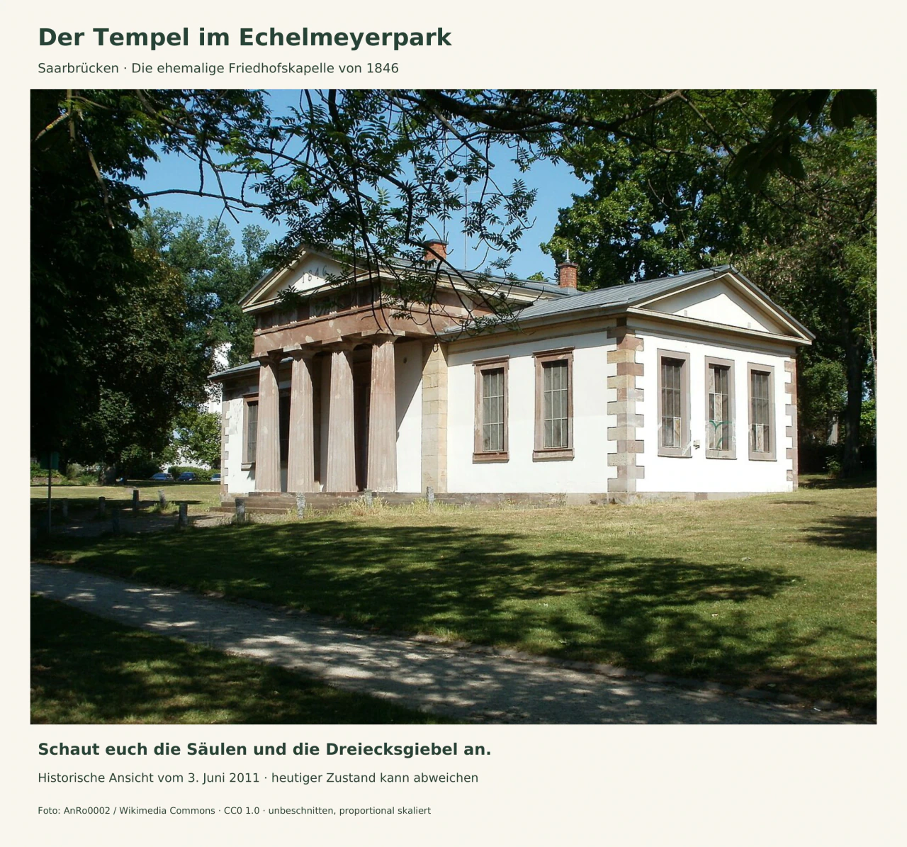 Die helle ehemalige Friedhofskapelle steht auf einer Wiese unter grünen Bäumen. Vier steinerne Säulen tragen den mittleren Dreiecksgiebel; rechts ist der seitliche Gebäudeflügel zu sehen.