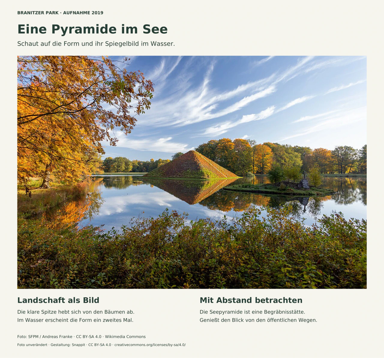 Eine bepflanzte Pyramide steht mitten im See und spiegelt sich fast vollständig im ruhigen Wasser. Goldgelbe Herbstbäume rahmen das linke Ufer; rechts liegt eine kleine Insel mit steinernem Kreuz. Sträucher füllen den Vordergrund.