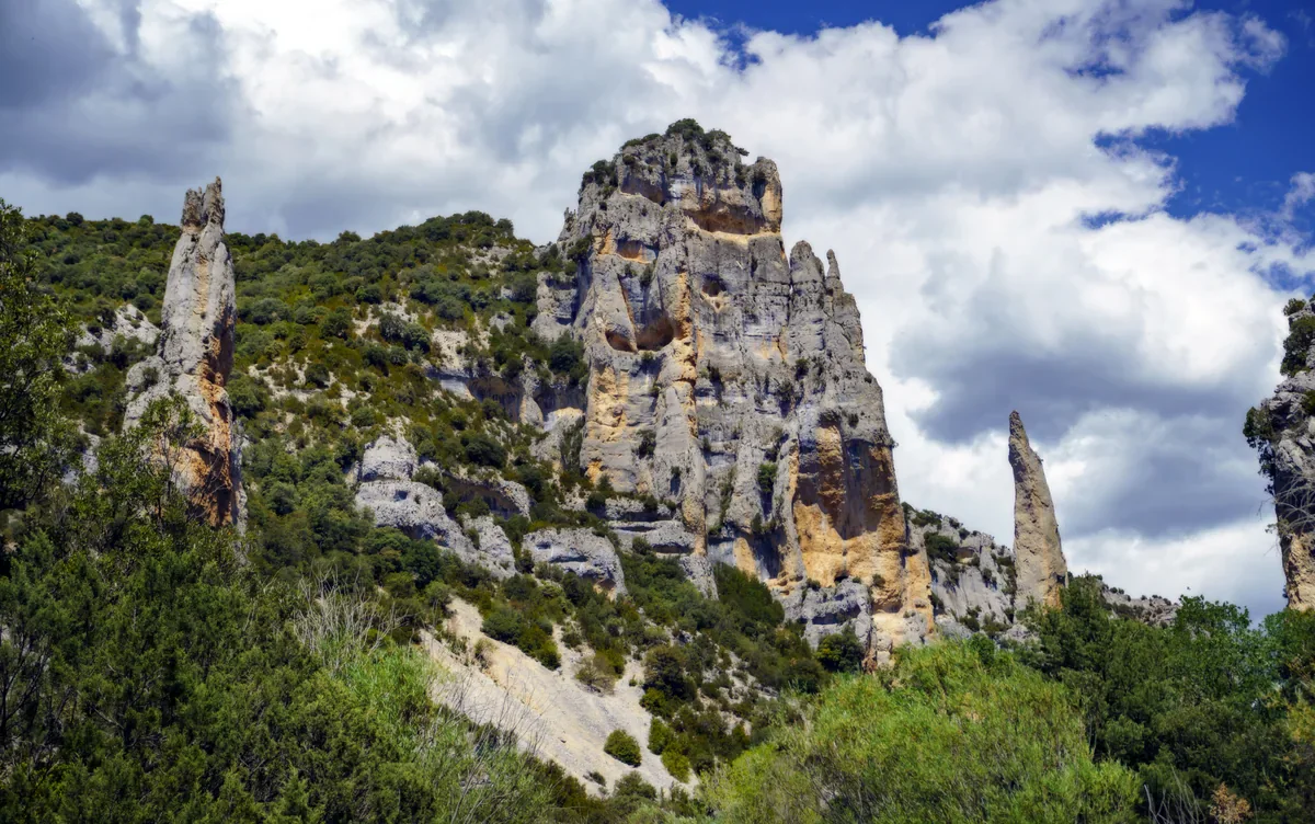 Tall pale limestone towers with hollows and narrow pinnacles rise above scrub-covered slopes beneath a blue sky and white clouds.