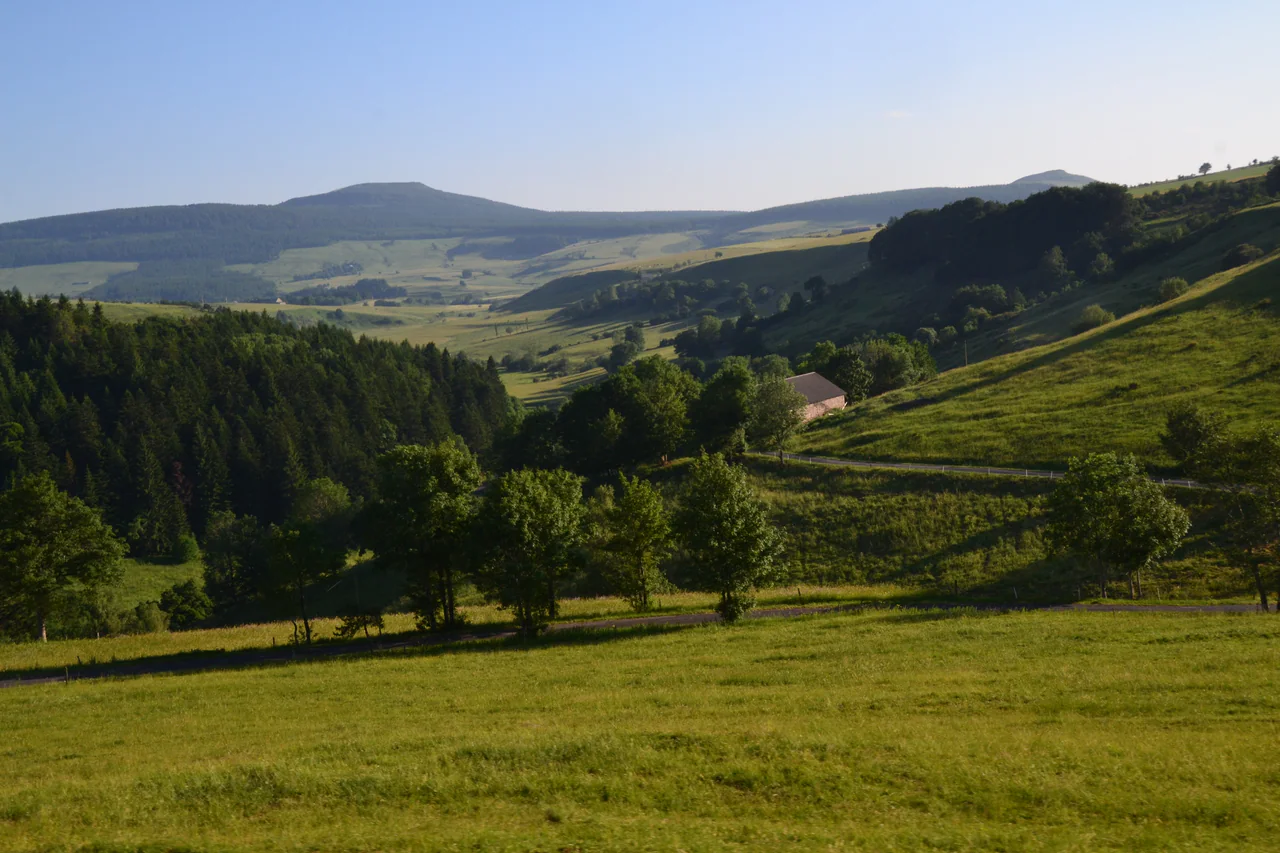 Green pastures and wooded slopes surround a farm building and winding roads below rounded uplands.
