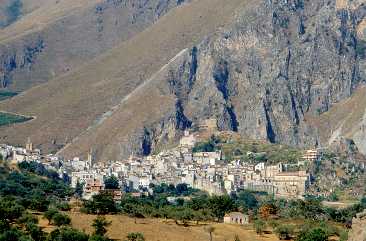 A compact town with church towers beneath a tall pale rock face, with scattered trees and fields in the foreground.