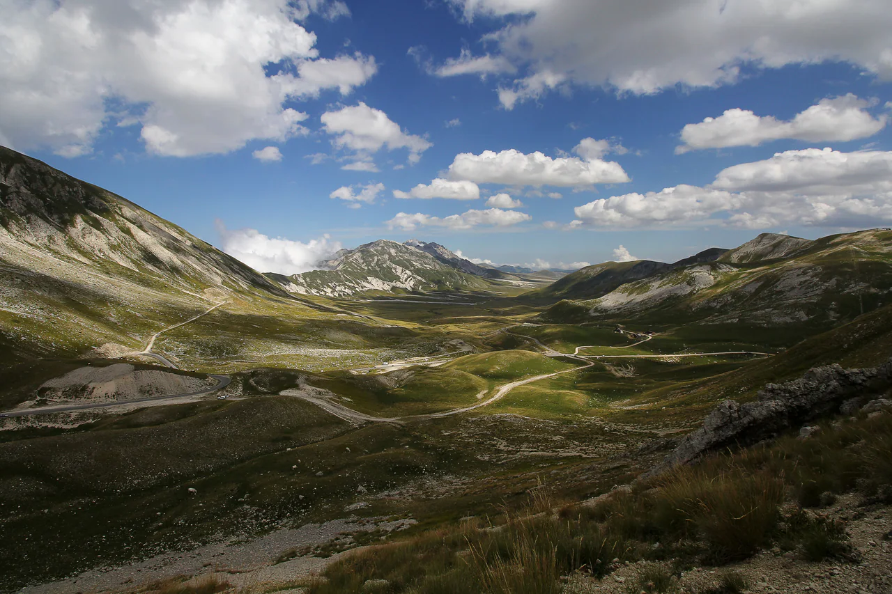 A broad grassy mountain valley between pale rocky slopes, with winding roads and a partly cloudy blue sky.