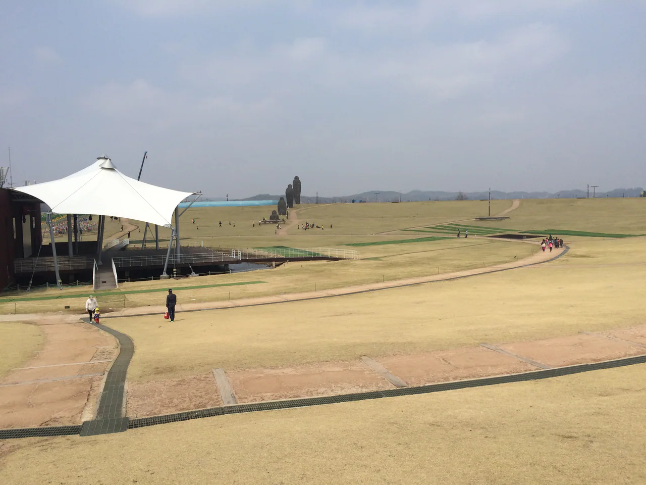 A broad pale lawn with scattered visitors, a white-roofed pavilion on the left and tall human-shaped sculptures in the distance.