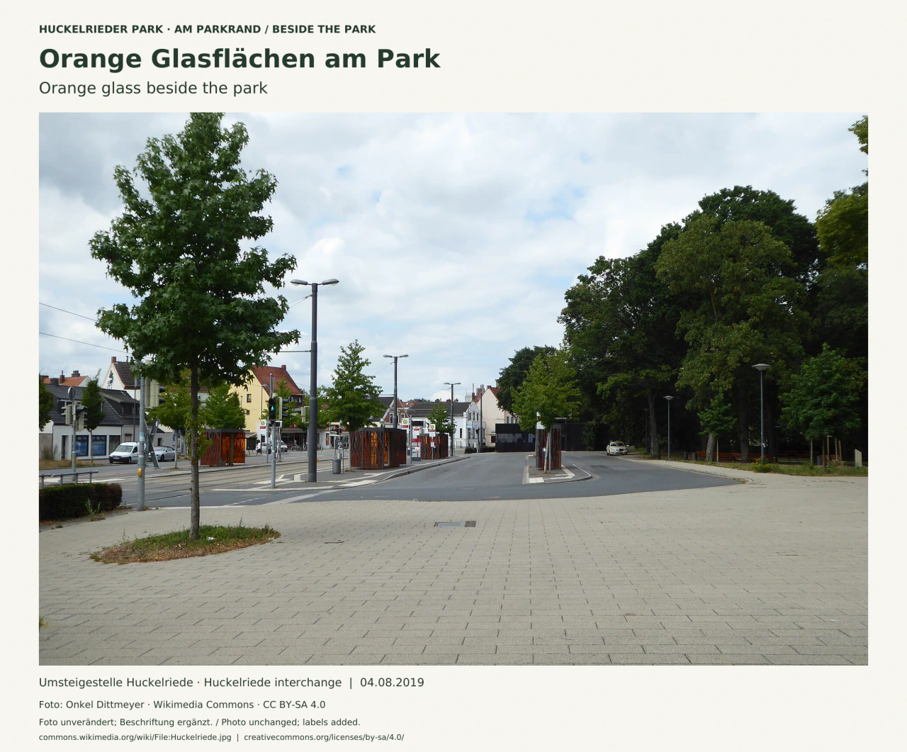 Breiter gepflasterter Bereich der Umsteigestelle Huckelriede mit orange verglasten Wartehäuschen vor grünen Bäumen; im Vordergrund sind Fahrspuren und Gehflächen zu sehen.