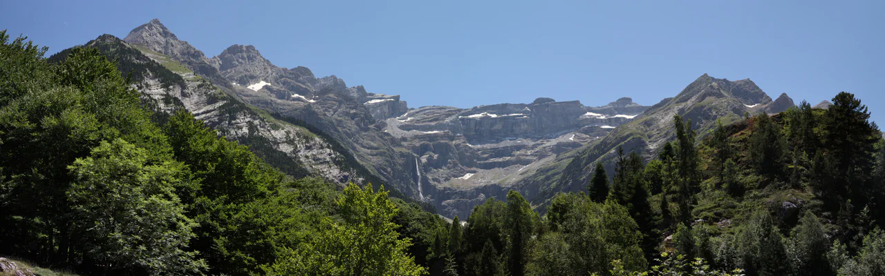 A wide arc of layered grey cliffs, a thin waterfall and patches of snow above green woodland under a clear blue sky.