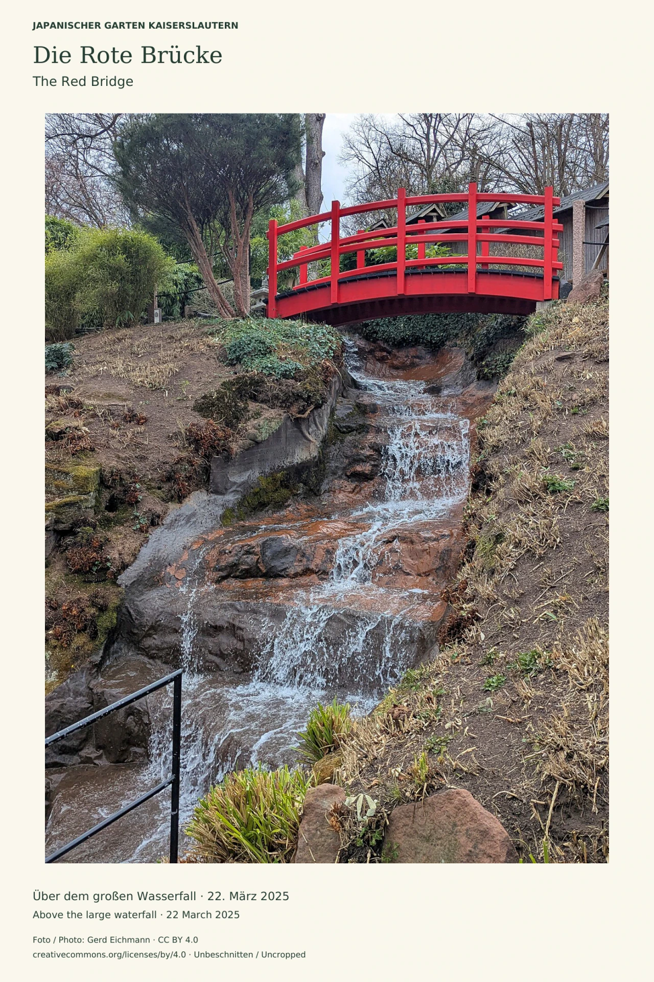Eine leuchtend rote, gebogene Holzbrücke überspannt einen Wasserfall über braunen Felsen. An den Hängen wachsen Pflanzen; dahinter stehen teils kahle Bäume. Links unten ist ein schwarzes Geländer sichtbar.