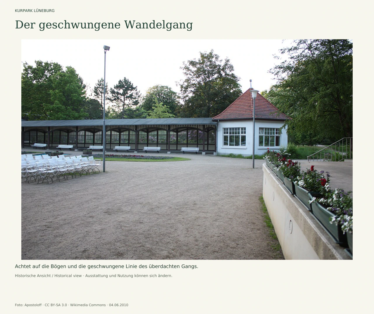 Ein überdachter, verglaster Wandelgang führt in einem Bogen zu einem weißen Pavillon mit rotem Dach. Weiße Bänke stehen davor, links befinden sich Stuhlreihen und rechts der bepflanzte Rand der Bühne.