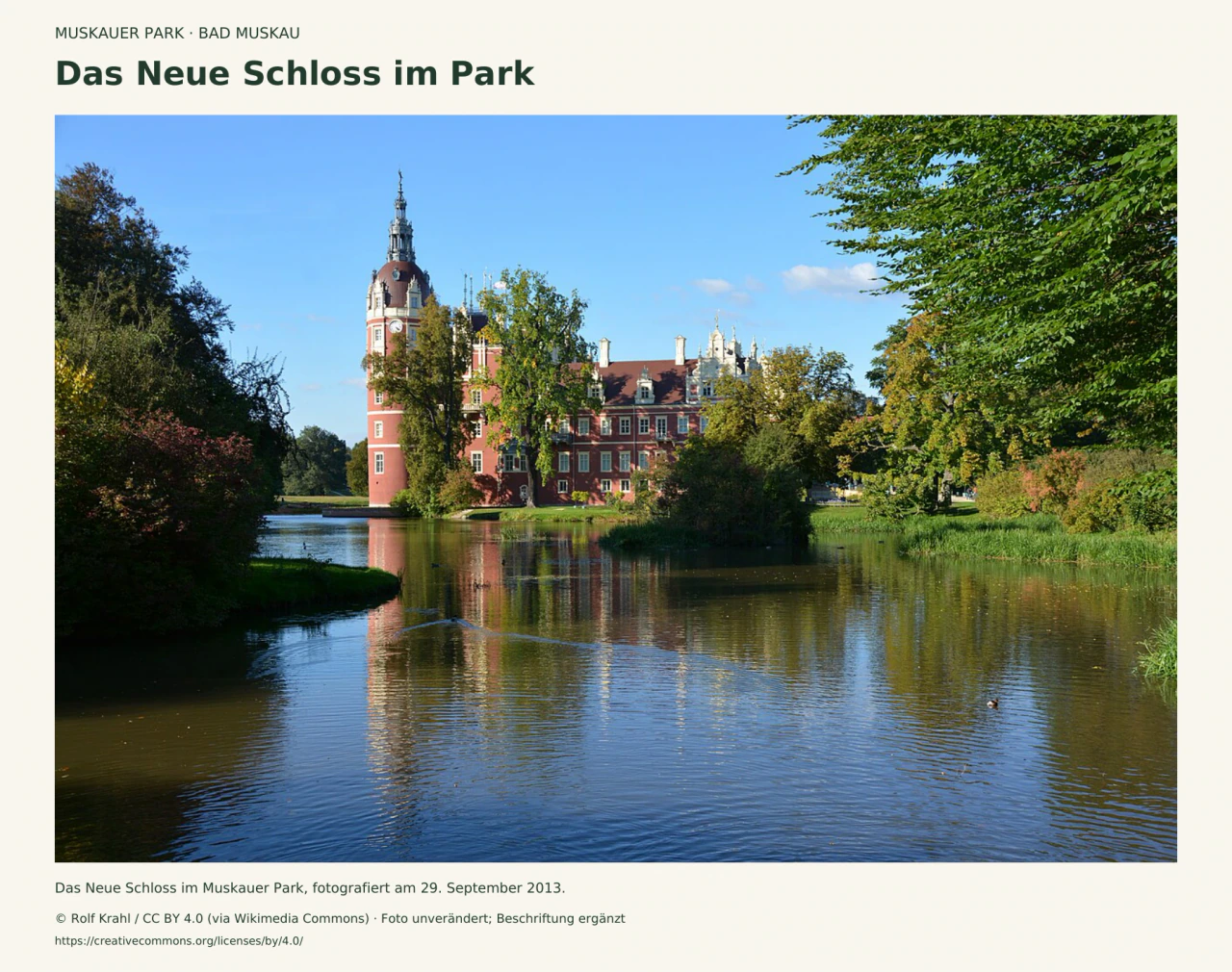 Das rötliche Neue Schloss mit hohem Turm spiegelt sich im Wasser; grüne Bäume rahmen die Ansicht im Muskauer Park, fotografiert im September 2013.