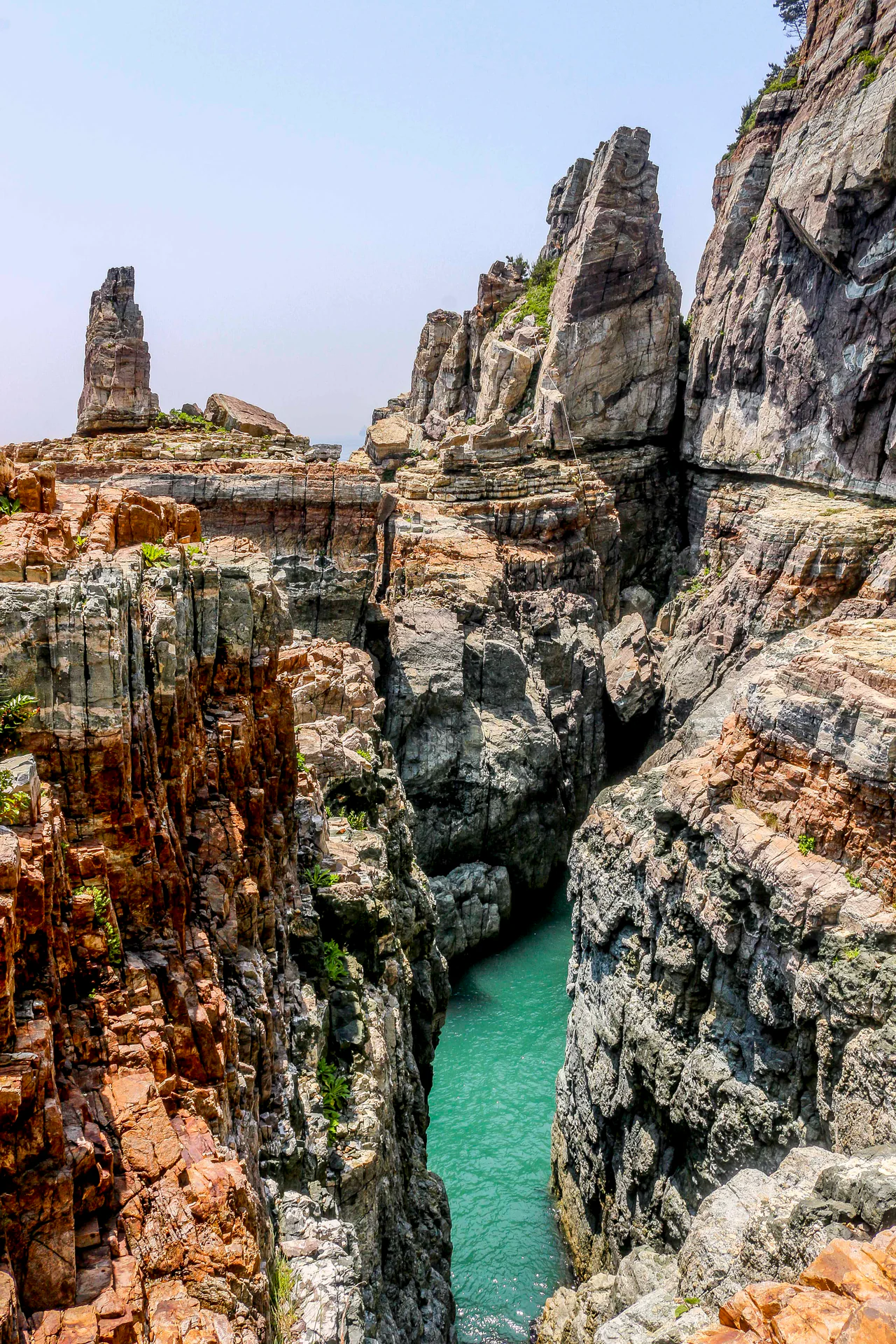 Tall layered rock faces and narrow stone spires surround a cleft of turquoise seawater, with sparse greenery along the upper rocks.