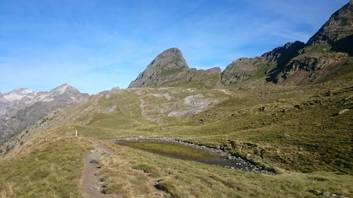An angular mountain peak rises above open grassy and rocky slopes, with a small shallow pool and a narrow footpath in the foreground.