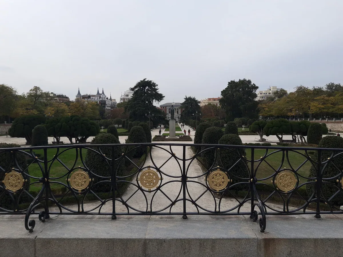 An ornamental black-and-gold railing overlooks symmetrical paths and rounded clipped trees, with distant visitors and city buildings under a grey sky.