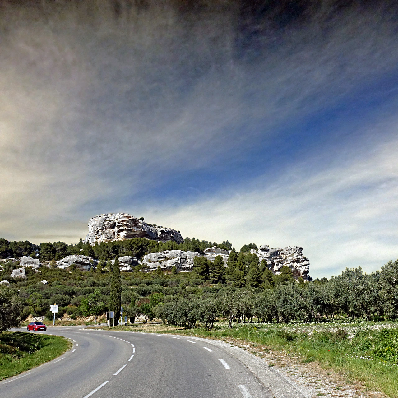 Pale limestone outcrops rise above dark trees and rows of olive trees; a curving road, a red car and signposts occupy the foreground beneath a strongly toned, streaked sky.