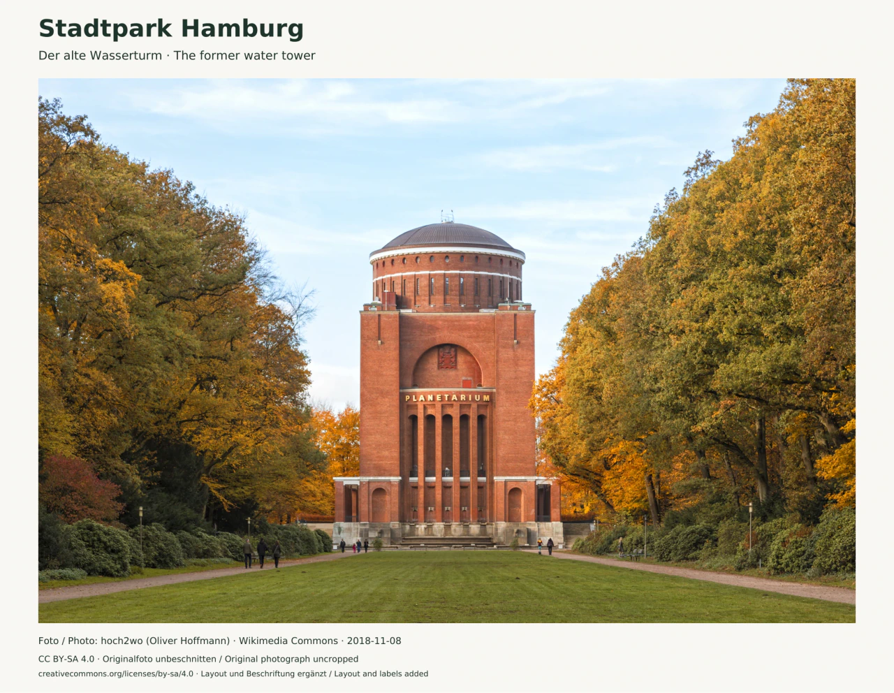Das rote Backsteingebäude des Hamburger Planetariums steht mittig zwischen goldgelben Herbstbäumen. Eine Rasenachse und zwei seitliche Wege mit einzelnen Spaziergängern führen darauf zu. Über den hohen Pfeilern steht PLANETARIUM.