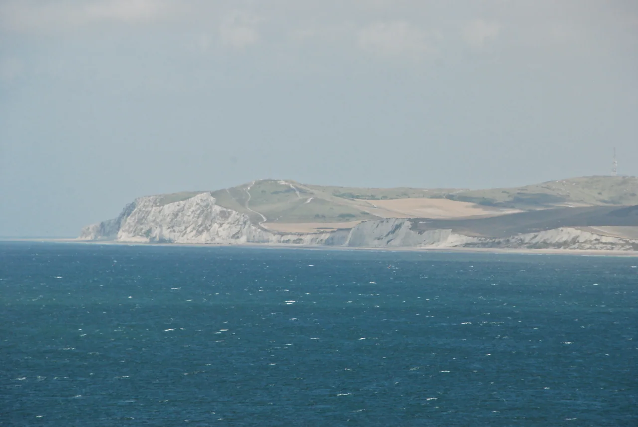 Pale chalk cliffs and fields across blue sea beneath a hazy grey sky, with a small mast on the distant ridge.