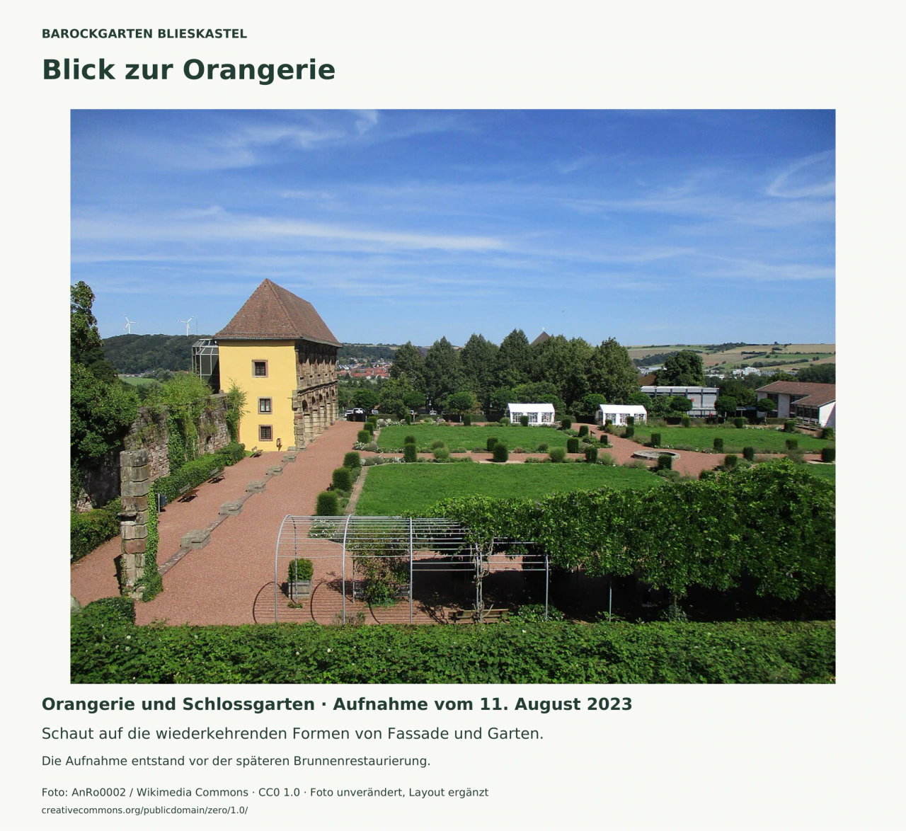 Blick von oben über Hecken, Rasenfelder und sich kreuzende Kieswege. Links steht die gelbe Seitenwand der Orangerie mit ihrem steinernen Bogenbau. Hinten sind zwei weiße Veranstaltungszelte und die Landschaft zu sehen.