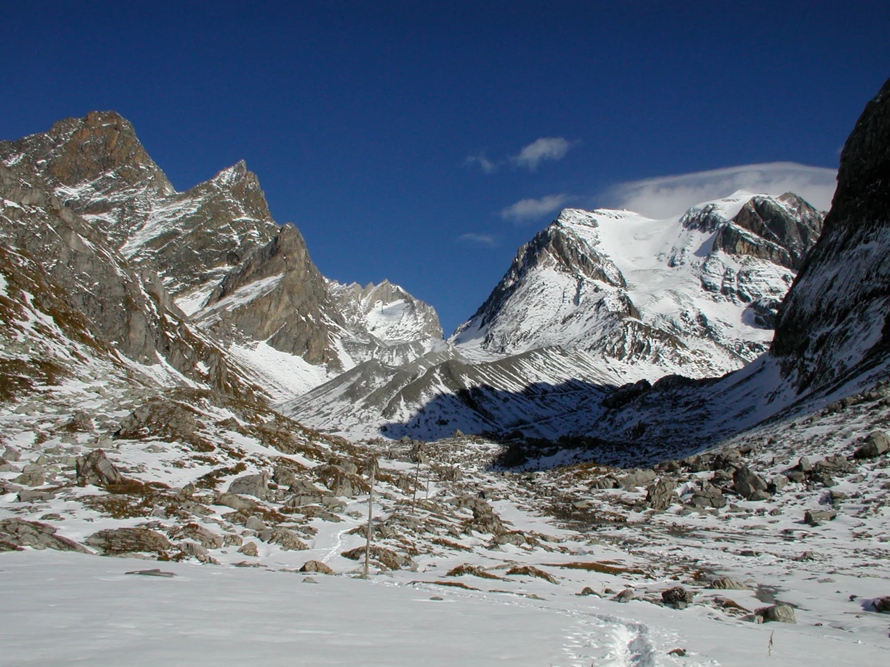 Snow covers a rocky Alpine basin below jagged ridges and a broad snowy mountain face under a deep blue sky; thin poles and tracks cross the foreground.