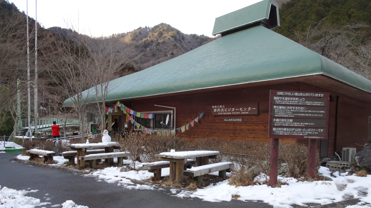 A low wooden visitor-centre building with a green roof, Japanese and English signs, snow-covered benches and wooded hills behind.