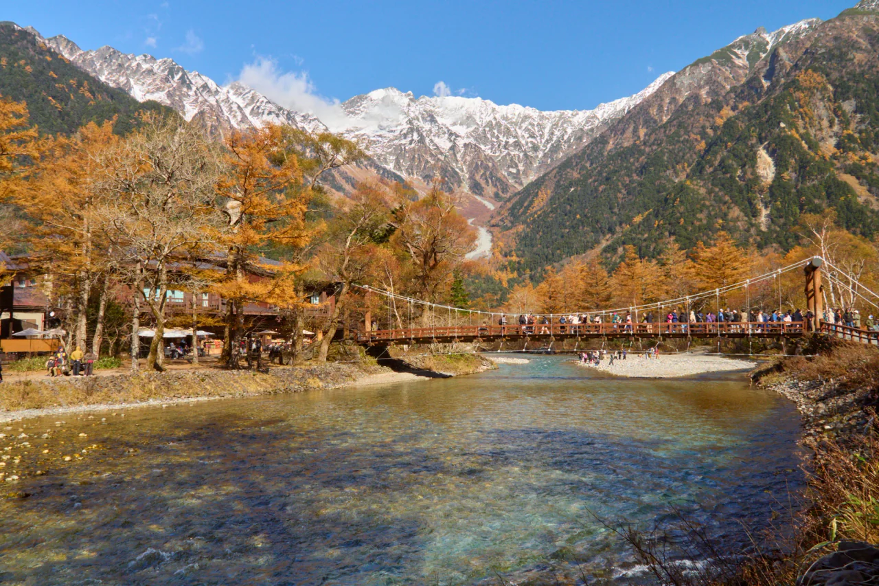 A wooden suspension bridge crowded with visitors spans a clear river, with golden autumn trees and snow-dusted mountain ridges behind.