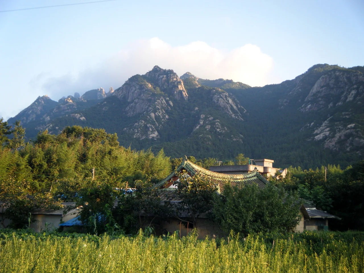 Forested granite ridges rise behind green fields, trees and low village rooftops beneath a pale blue sky.