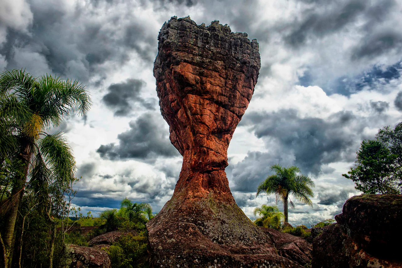 A tall reddish sandstone formation with a narrow lower section, surrounded by green vegetation beneath a cloudy sky.