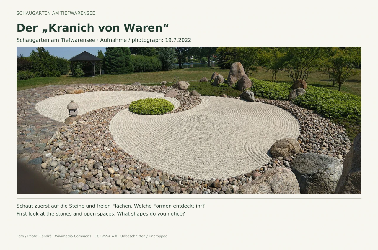 Weiße Kiesflächen mit gebogenen Rechenspuren, eingerahmt von runden Steinen und größeren Felsblöcken. Links steht eine kleine Steinlaterne; dahinter sind Rasen, Gehölze und ein Pavillon zu sehen.