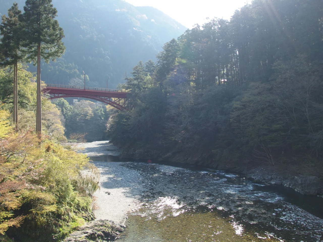 森に囲まれた渓谷の川と白っぽい砂利の岸に日が差している。木々の間には赤い橋が架かり、奥にはかすんだ森林の山並みが見える。