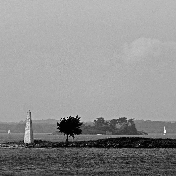 Black-and-white view across water to a low islet with a stone beacon and tree, with wooded shores and small sails beyond.