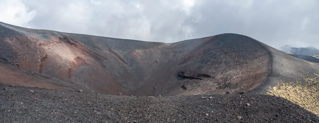 Un ampio cratere di rocce vulcaniche grigio scuro e rossastre sotto nuvole basse, con rada vegetazione chiara lungo il bordo destro.