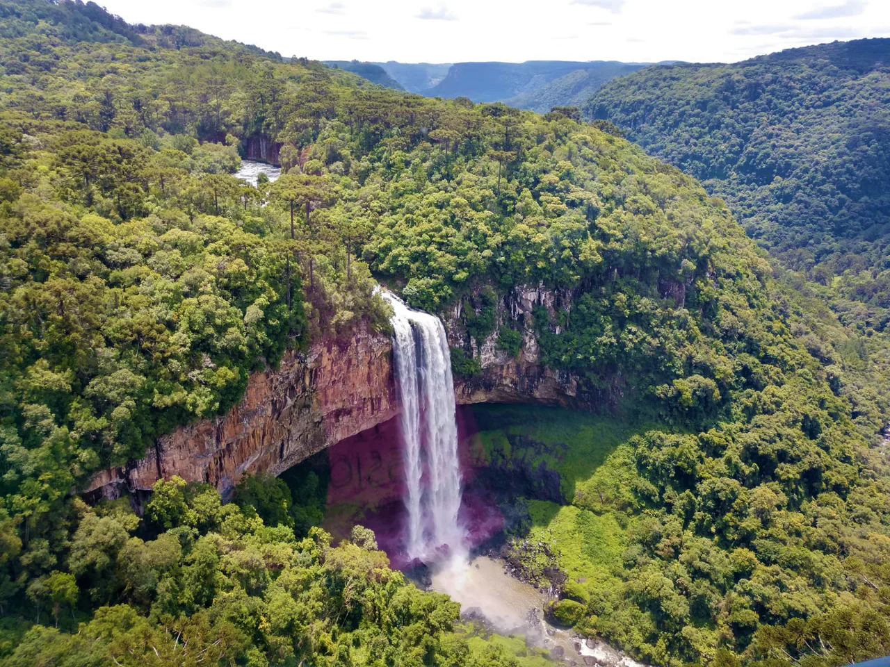 A broad white waterfall drops over a curved rock wall into a wooded valley, with dense green forest above and around the cascade.