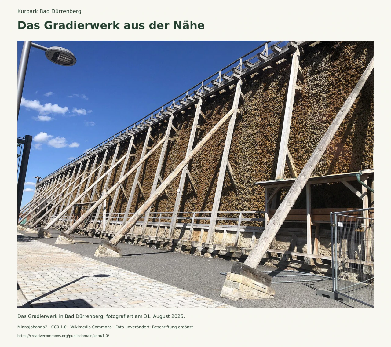Das hohe Gradierwerk zieht sich diagonal ins Bild: helle Holzstützen tragen eine dichte braune Reisigwand; davor liegen ein Geländer und ein befestigter Weg.