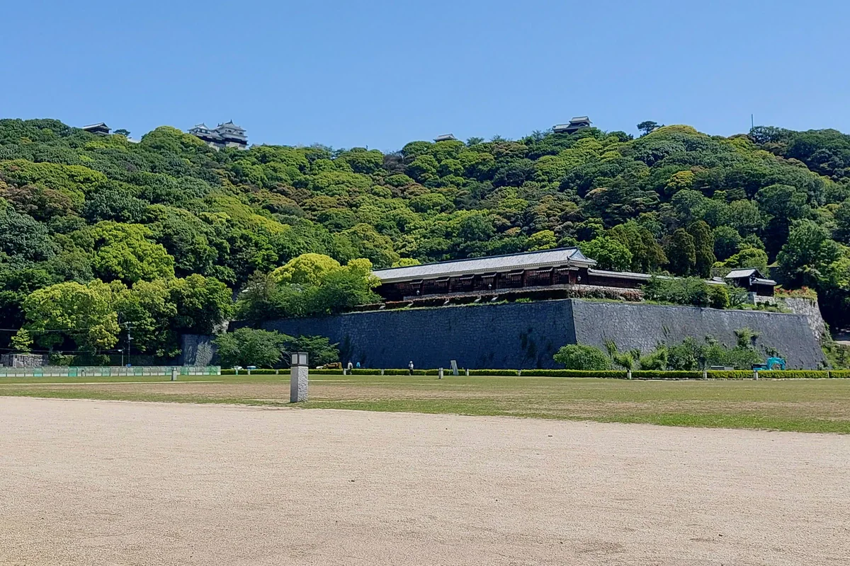 An open gravel-and-grass foreground leads toward stone walls and a densely wooded hill, with castle roofs visible above the trees.