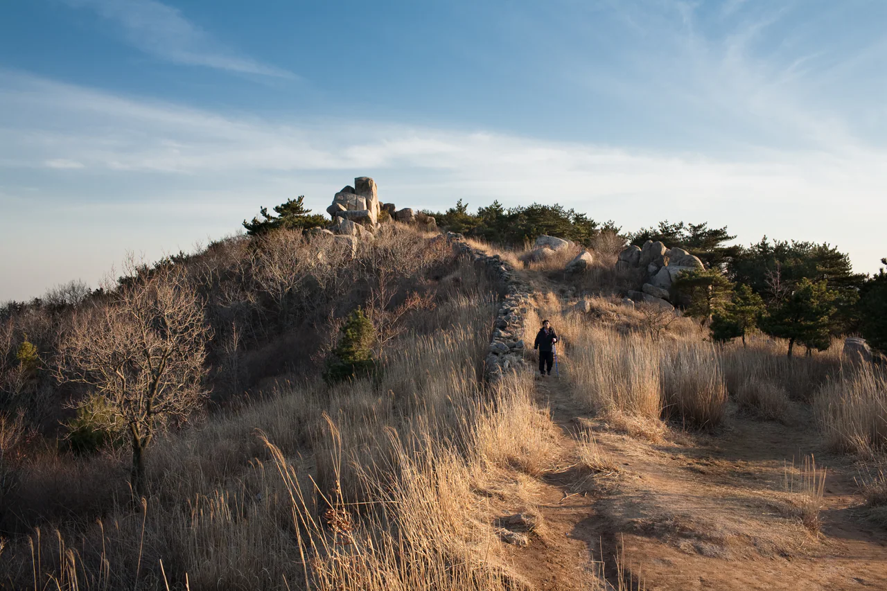 A low stone wall runs through dry grass past granite boulders and pines, with a small hiker on the adjacent path.