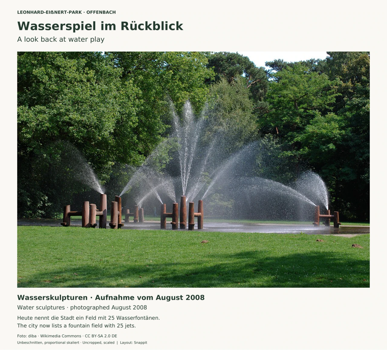 Mehrere braune, aufrecht stehende Wasserskulpturen mit kurzen seitlichen Verbindungen sprühen hohe Wasserbögen vor dicht belaubten Bäumen; im Vordergrund liegt Rasen.