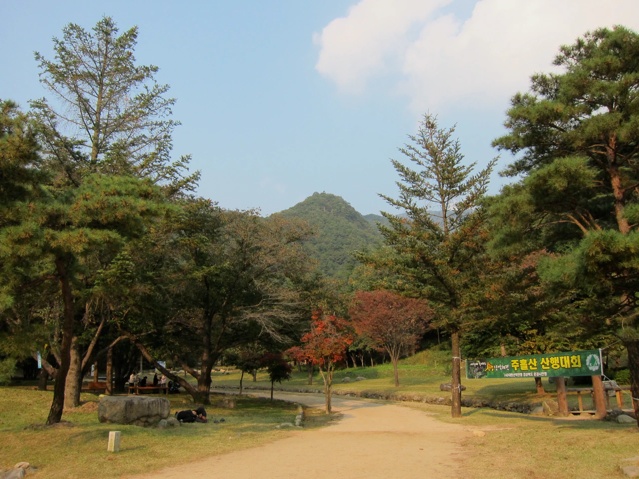 A broad dirt path curves between pine and autumn-colored trees toward a wooded ridge, with a few resting visitors and a Korean hiking-event banner at the right.