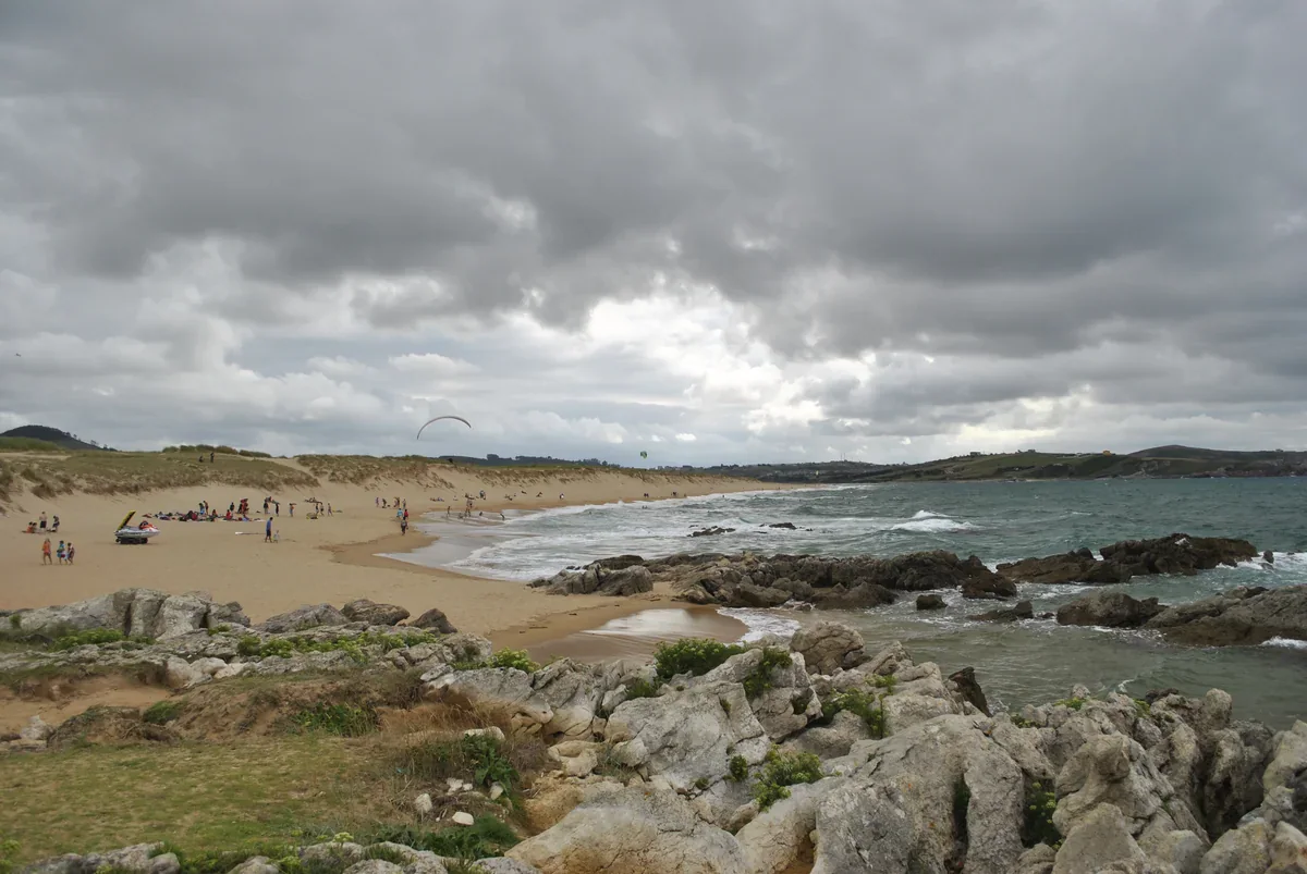 Dunes rise behind Valdearenas beach, with small distant beachgoers, breaking waves and rocks in the foreground beneath a cloudy sky.