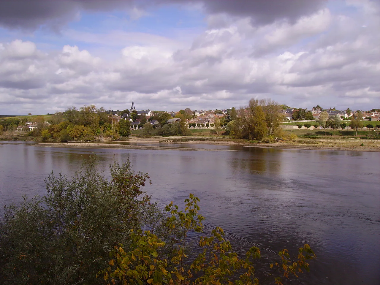 A broad river, sandy margin and tree-lined village beneath a cloudy sky.