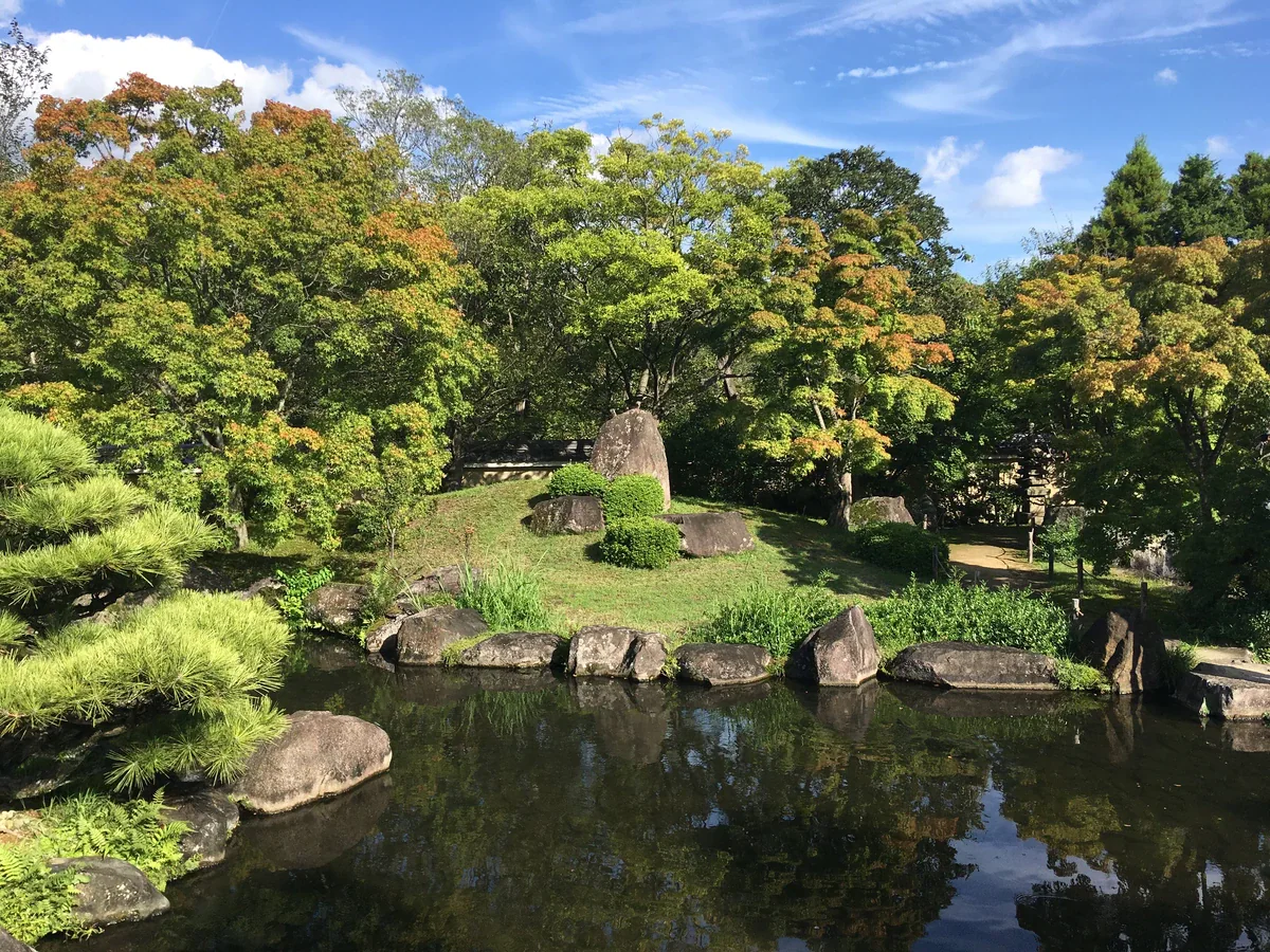 Rocks and clipped shrubs form a planted mound beside a reflective pond, backed by leafy trees at Koko-en.