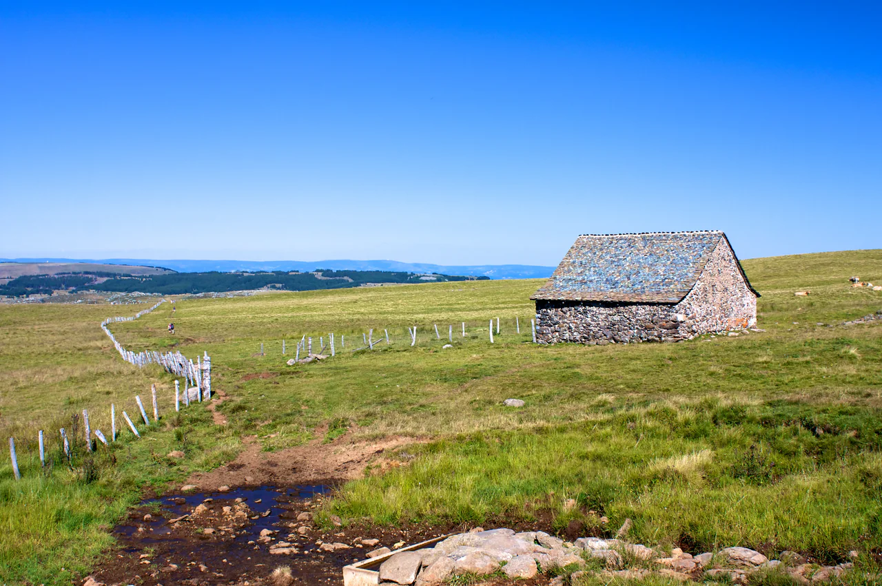 A low stone buron with a broad stone roof stands in open pasture beside a fence; a muddy, stony track crosses the foreground, with distant walkers and cattle under blue sky.
