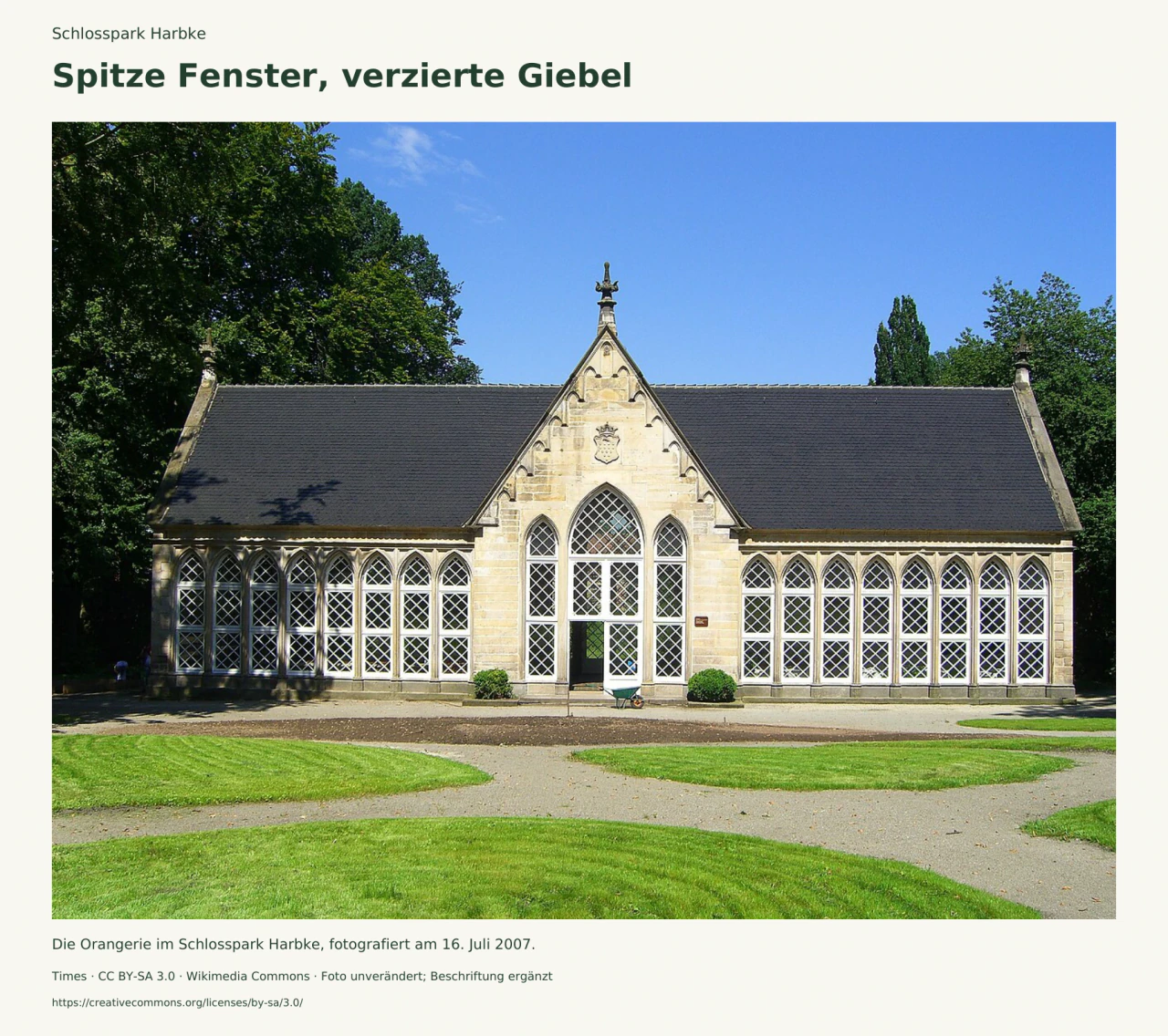 Die helle Orangerie Harbke mit einer Reihe spitzbogiger Fenster, weißem Rautengitter und hohem Mittelgiebel; davor liegen Rasenflächen und Wege.