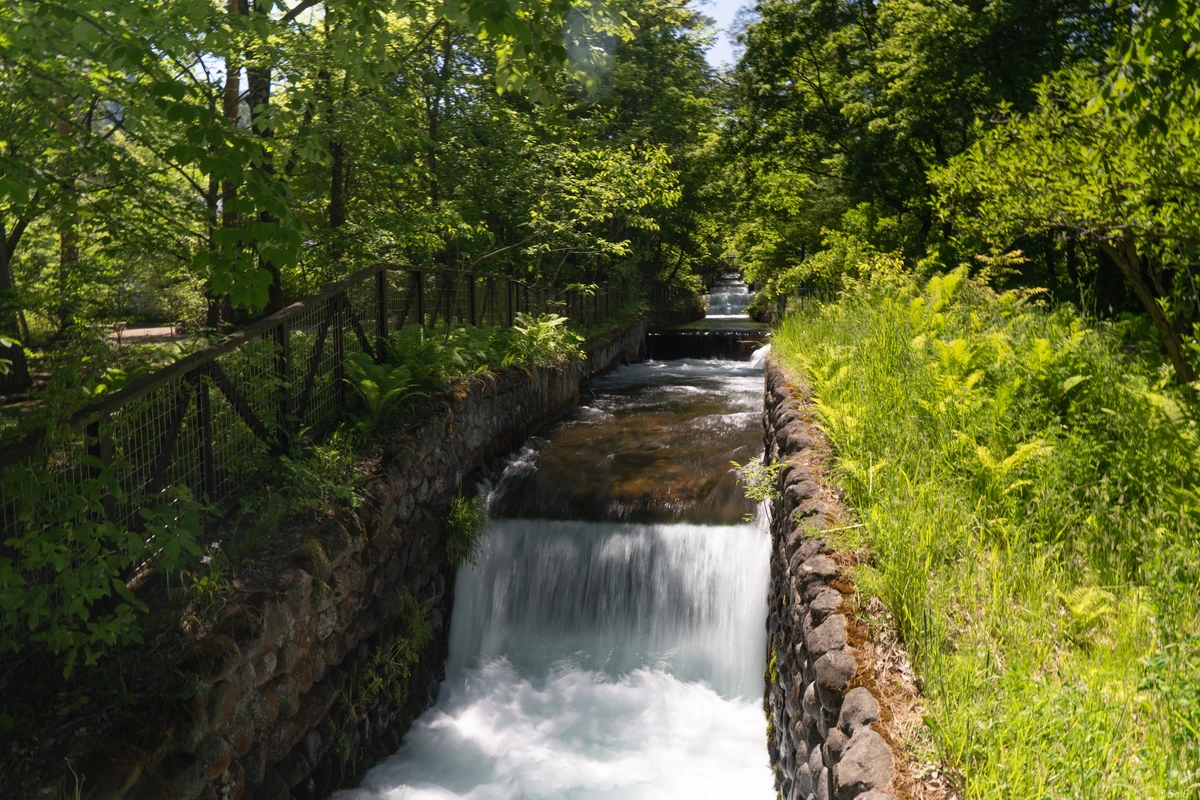 Water cascades over low steps in a stone-lined channel surrounded by green trees; a fence separates it from the path.