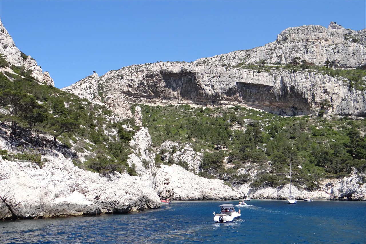 Pale limestone cliffs and green scrub above deep-blue water at Morgiou, with several small boats in the foreground.