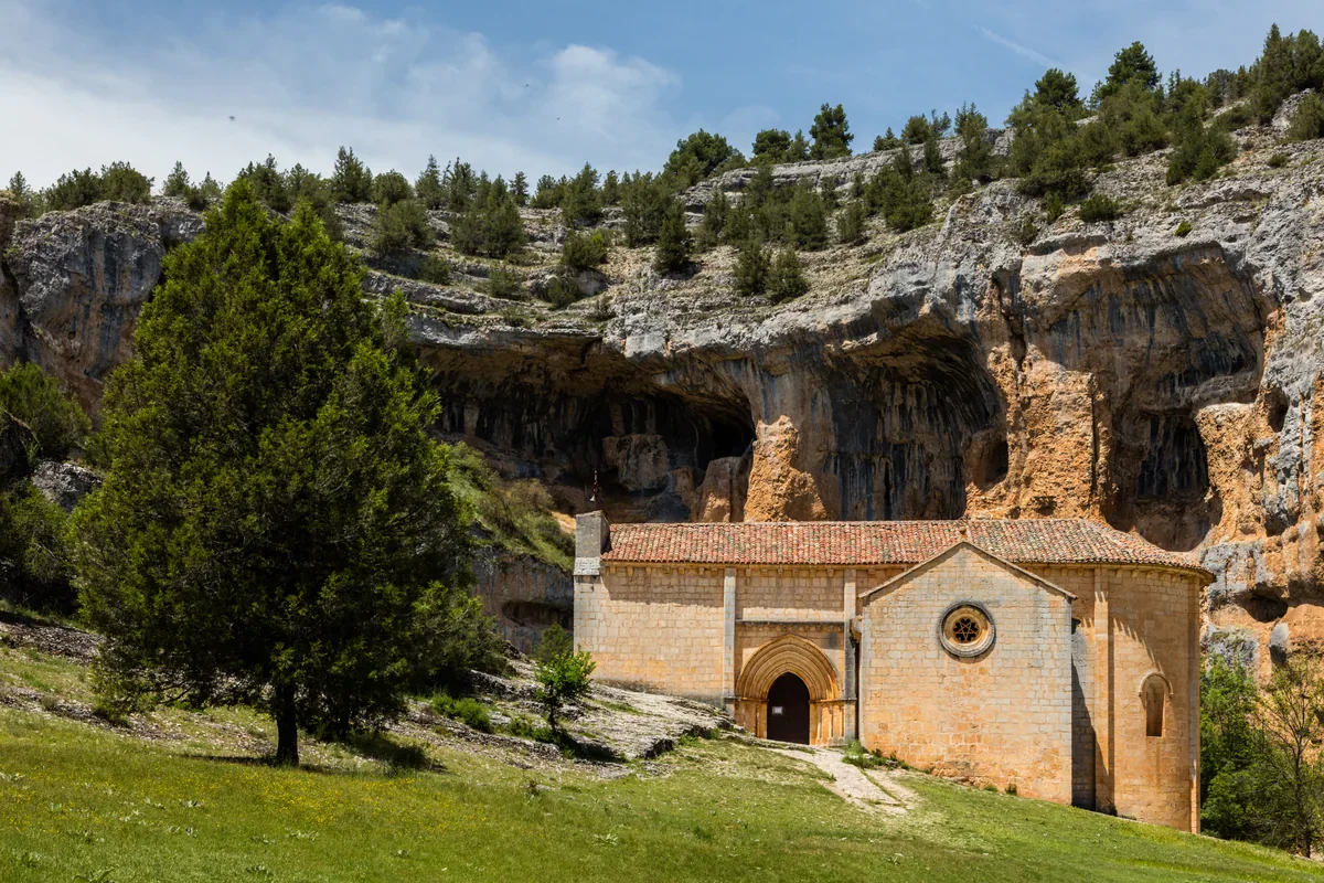 A pale stone hermitage with a tiled roof and round window sits below an orange-grey cliff, with grass and a dark green tree in the foreground.