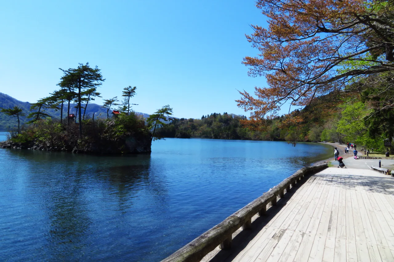 A wooden lakeside walkway curves beside blue water, with a small tree-covered rocky island on the left and distant walkers along the wooded shore on the right.