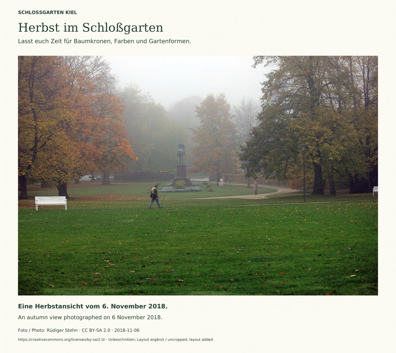 Blick über eine Rasenfläche im Kieler Schloßgarten auf das Kaiser-Wilhelm-Reiterdenkmal im Herbstnebel; farbige Bäume, geschwungene Wege, weiße Bänke und zwei kleine Besucherfiguren.