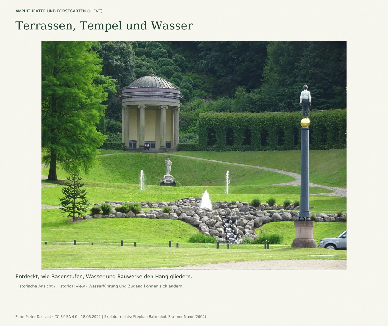 Terrassierter grüner Hang mit Wasserfontänen und einem steinernen Wasserlauf. Ein runder Säulentempel steht oben links, eine helle Statue in der Mitte; rechts steht eine Männerfigur auf einer hohen dunklen Säule.