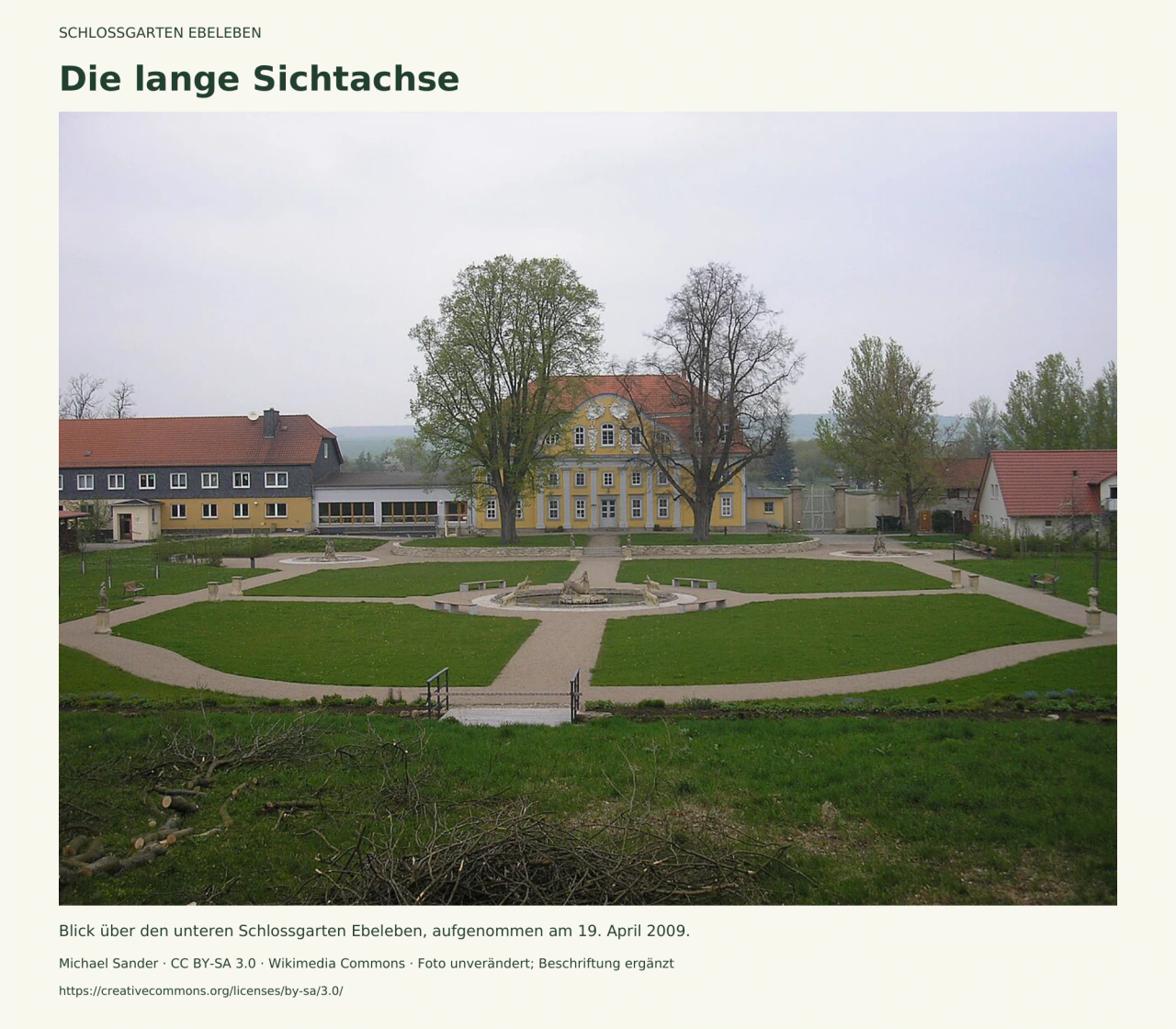 Symmetrische Kieswege und Rasenflächen führen an einem runden Figurenbrunnen vorbei zur gelben ehemaligen Orangerie; Bäume und weitere Gebäude rahmen die Anlage, im Vordergrund liegen geschnittene Äste.