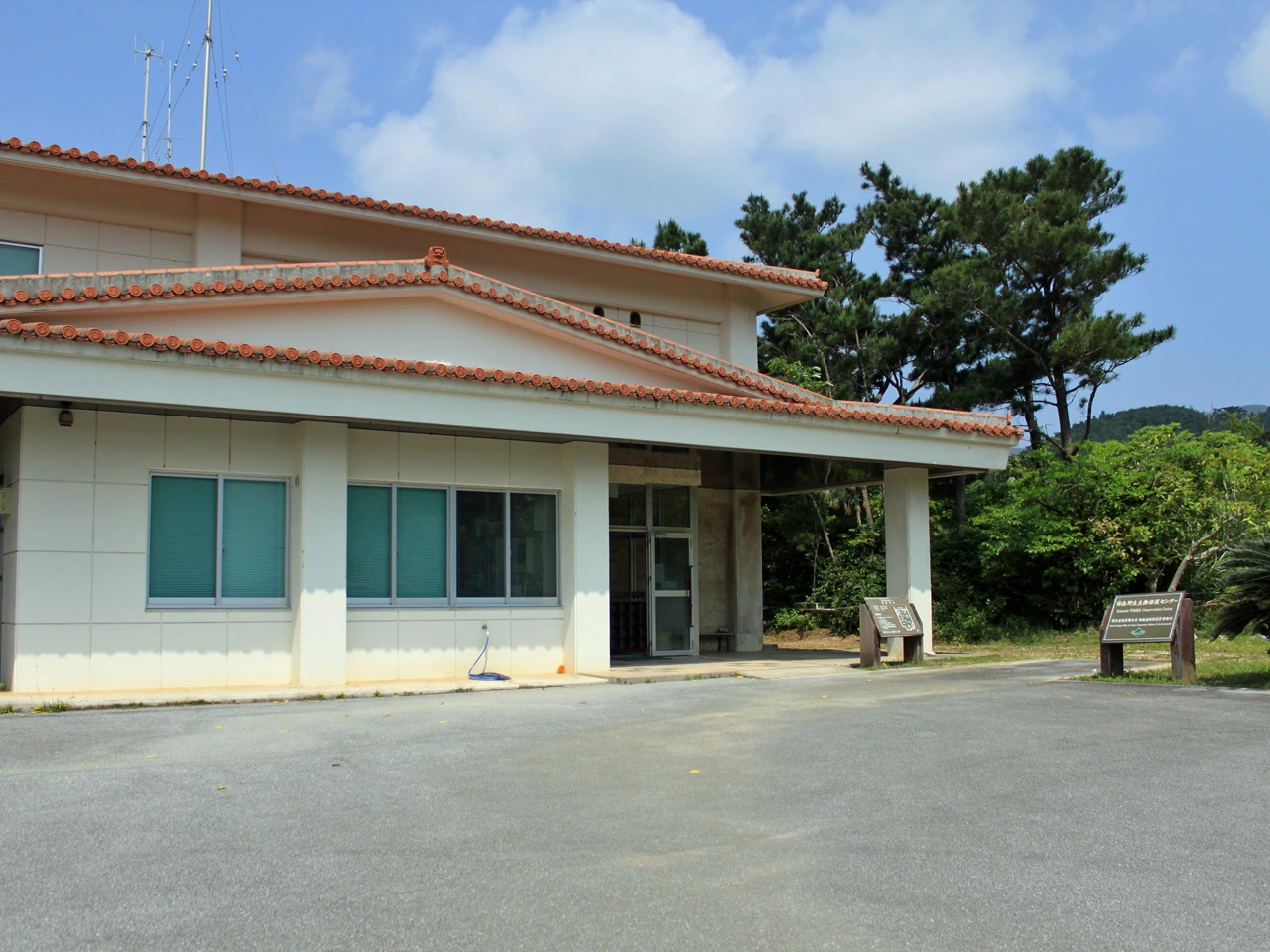 A low cream-coloured building with red roof tiles and a shaded entrance beside trees and a paved forecourt.
